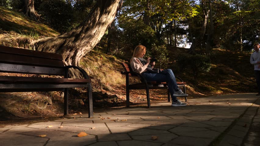 4k, slow-motion, girl with a camera sits on a bench in a picturesque place. Passers pass by.