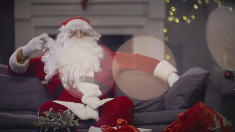 Santa Claus touching beard and waiting Christmas night for celebration and congratulation. Santa Claus sitting on couch on background Christmas present box pending holiday