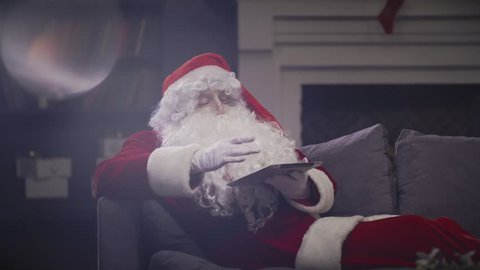 Santa Claus using tablet sitting on couch on background Christmas fireplace. Santa Claus looking tablet at Christmas eve for communication with children