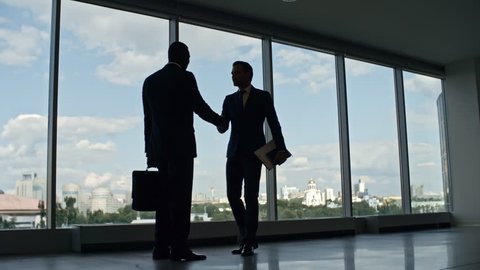 Silhouettes of business partners greeting each other with handshake and walking together through corridor in office building with panoramic windows