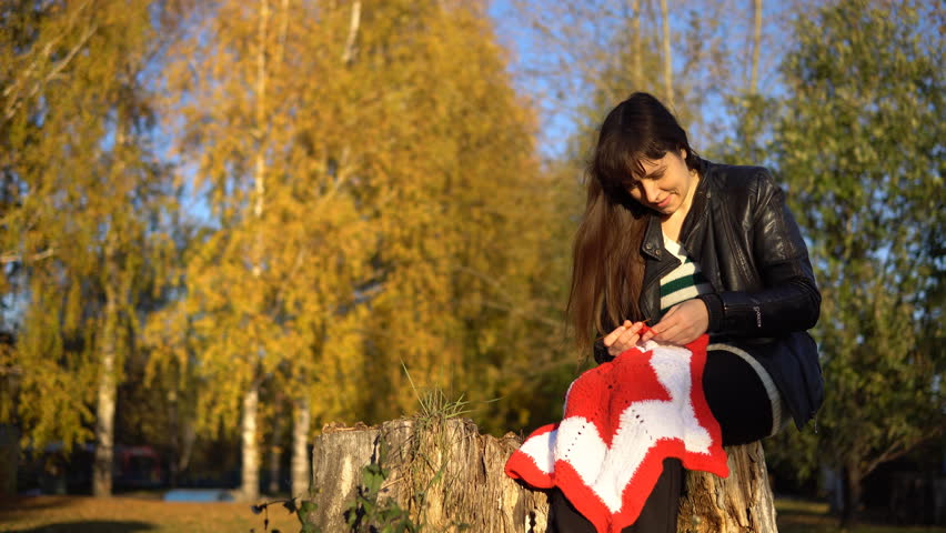 Young brunette woman in a striped blouse and a black leather jacket crochet knit red-and-white plaid, sitting in the autumn or winter park