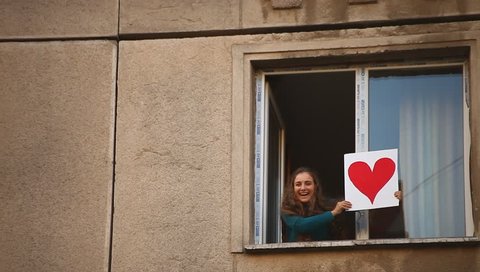 Cheerful young adult woman shows a read heart valentine into the window.
 Vídeo Stock