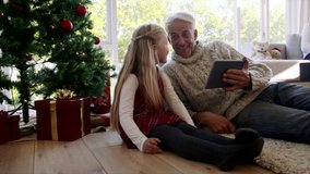 Smiling little girl and grandfather sitting on floor by christmas tree with digital tablet, with grandmother sitting on sofa at the back. Two generation family at home during christmas festival.