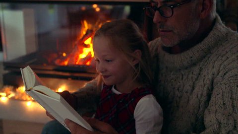 Senior man reading a book to little girl near fireplace. Senior man with grandaughter reading a story book.