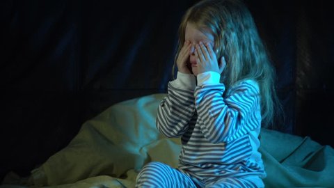 A upset little girl in striped pajamas sits on the bed at night in her room and cries covering her face with her hands, then wipes away her tears and tries to calm down.