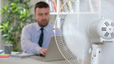 The video is about a sweaty man employee trying to refresh himself because of summer heat with a fan.Shot focus is fixed on the fan ventilator and the businessman acts out of focus on the background