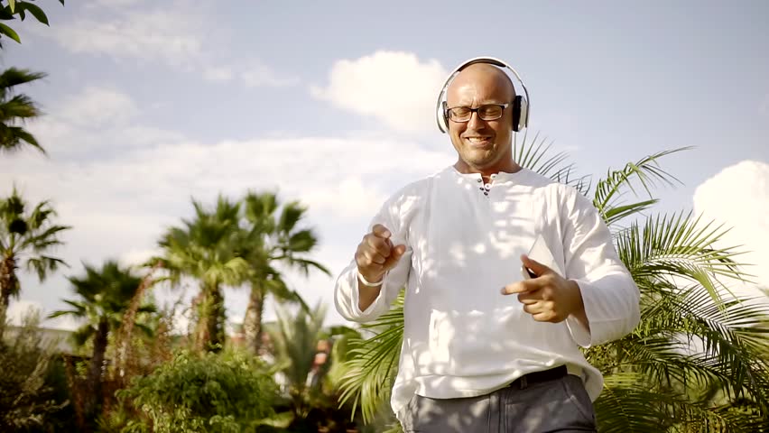 middle-aged man is listening to a music by headphones and dancing in yard near palms in sunny day