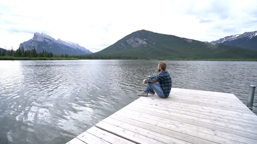 View of a young man sitting on a wooden lake pier contemplates the beautiful mountain view, Banff national park, Canada