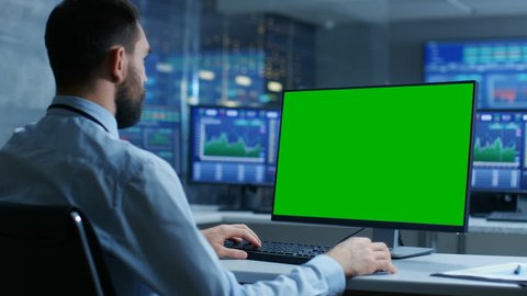 Over the Shoulder View of Stock Market Trader Working on a Computer with Isolated Mock-up Green Screen. In the Background Monitors Show Stock Ticker Numbers and Graphs. Shot on RED EPIC-W 8K Camera.
