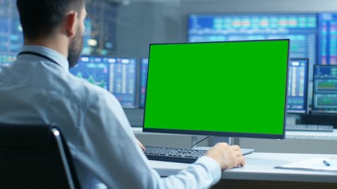 Over the Shoulder View of Stock Market Trader Working on a Computer with Isolated Mock-up Green Screen. In the Background Monitors Show Stock Ticker Numbers and Graphs. Shot on RED EPIC-W 8K Camera.