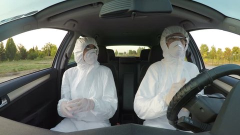 Young funny scientists sitting in car dressed in hazmat wear dancing and feeling energized and celebrating achievement