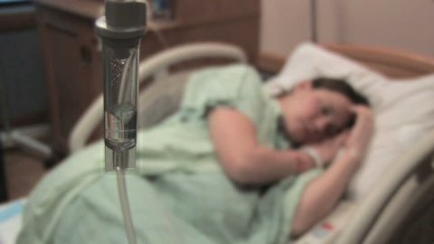 A sick woman lays in a hospital bed behind an IV drip chamber.