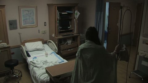A woman in a hospital gown walks with her IV across the hospital room and sits down on the bed