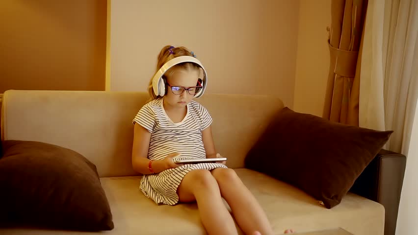 youngling girl is sitting in a sofa, she is wearing glasses and wireless headphones, watching films