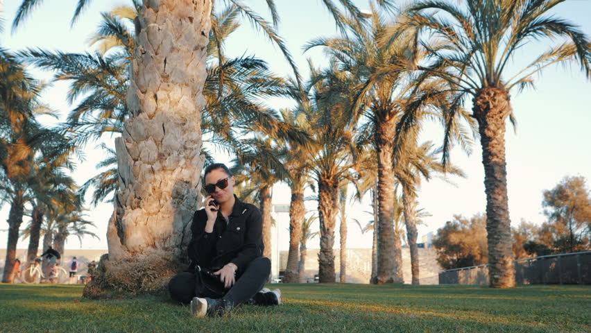 Young caucasian brunette trendy woman in sunglasses talking on cell phone and smiling while sitting under the palm tree. Travel concept. Slow motion