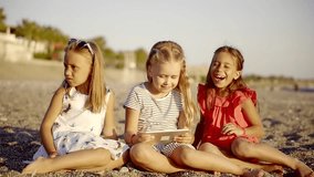 cute little girlies are watching video in an application of a tablet, sitting on an empty beach