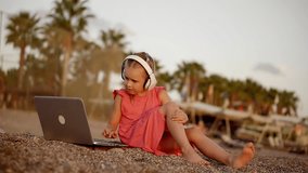 little girl is watching cartoons by laptop and listening sounds by headphones, sitting on a coast