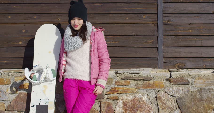 Beautiful young woman in pink skiwear and knitted cowl standing with her snowboard near wooden and stone building wall at ski resort.