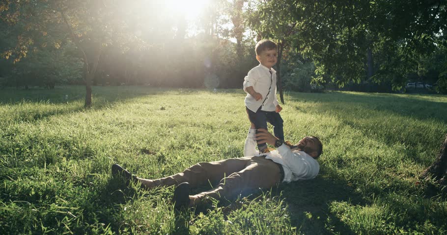 Young father and his little son are playing on the backyard. They are lying on the lawn togehter.