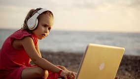 little girl is sitting on a beach in evening and using laptop, she is wearing wireless headphones