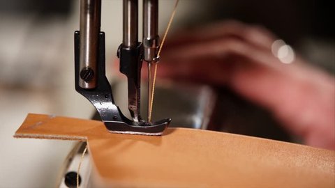 Industrial sewing machine is stitching detail of leather bag. Tailors hands are controlling process, needle is accelerating and slowing down, macro