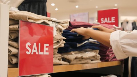 Female Hands Choosing Jeans from Stack in Clothing Store. Big Red Sale Sign in Shopping Mall. Black Friday Concept. 4K. ஸ்டாக் வீடியோ