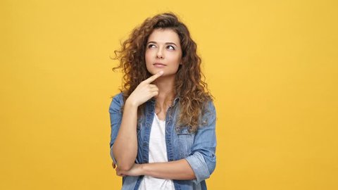 Smiling pensive curly woman in denim shirt having idea and looking at the camera with crossed arms over yellow background