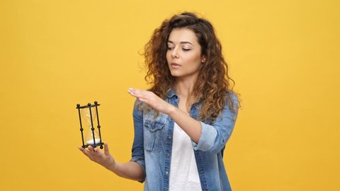 Young curly woman in denim shirt turns the hourglass and pointing at camera over yellow background