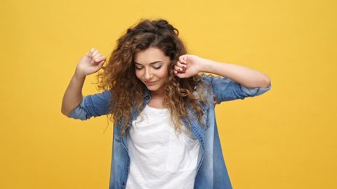 Happy carefree curly woman in denim shirt dancing and looking at the camera over yellow background
