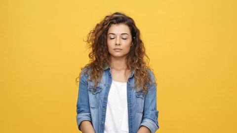 Tired bored curly woman in denim shirt touching face and looking at the camera over yellow background
