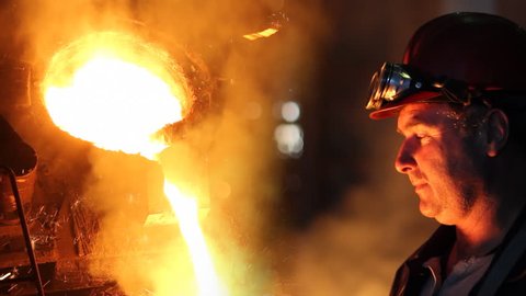 Hard work in the foundry, worker watching and controlling iron smelting in furnaces, too hot and smoky workplace. He puts on safety goggles