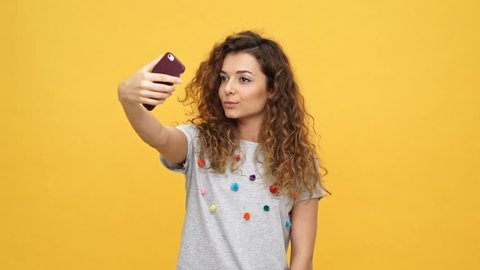 Smiling happy curly woman in t-shirt making selfie on smartphone over yellow background