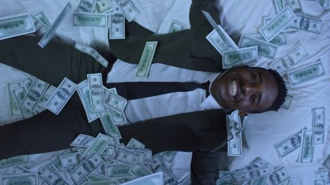Directly above shot of African-American businessman in suit sleeping in bed, then waking up and smiling with happiness as money raining on him from above