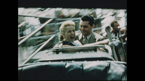 1950s: Man and woman sit in car of ride, spin around, smile. Amusement park ride spins. Parachutes rise and fall from high tower.