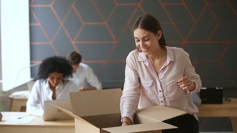 First day at new job concept, young happy woman unpacking box with her belongings standing near office desk, just hired smiling intern starting work in big company, moving into coworking space
