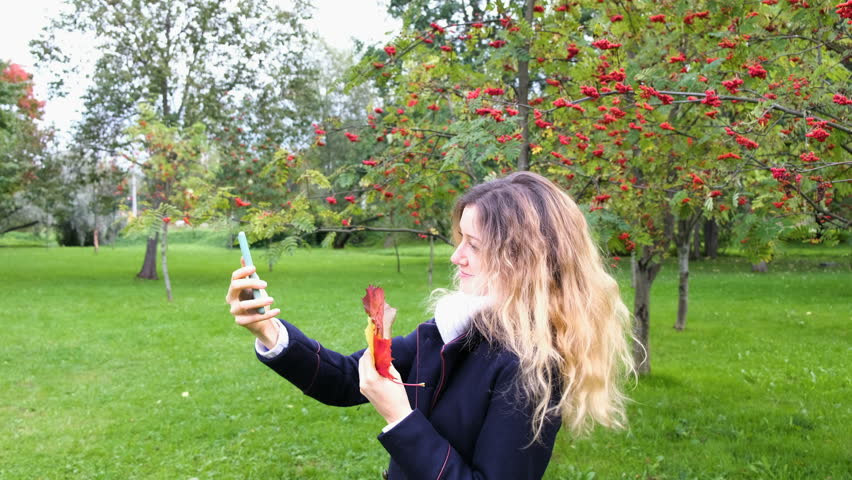 Cute young girl in Park taking a selfie on smartphone with maple leaves, 4K