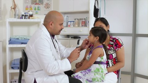 An Indian pediatrician or family doctor uses an otoscope to examine the eyes of a little girl who is being accompanied by her mother.