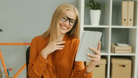 Attractive blonde young woman wearing glasses video chatting on her tablet device and waving hands. The blurred office background. Indoor.