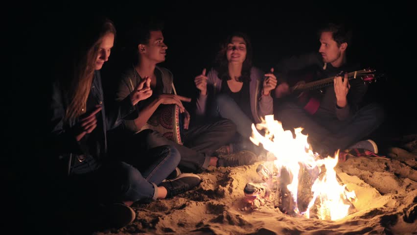 Two young women and two men sitting by the bonfire late at night and singing songs, playing guitar and percussion