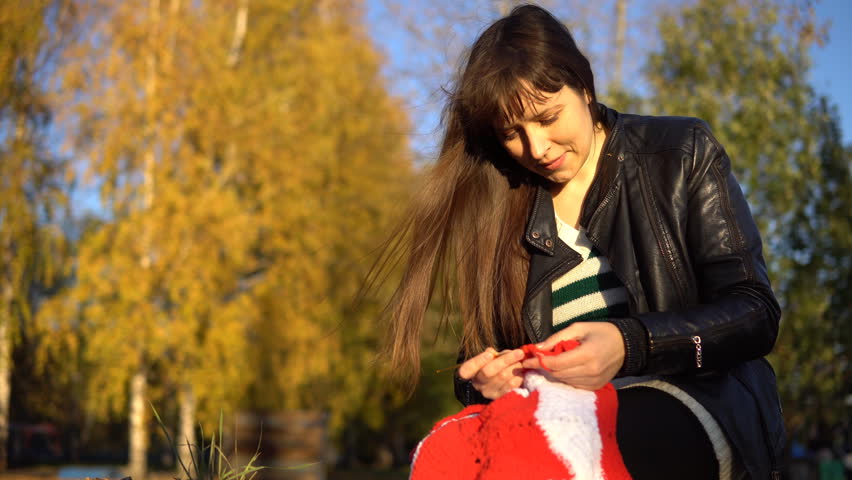 Young brunette woman in a striped blouse and a black leather jacket crochet knit red-and-white plaid, sitting in the autumn or winter park
