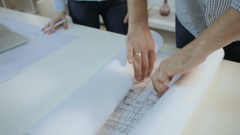 Two young designers unfold the drawings of products on a large table in the office-studio. in slow motion