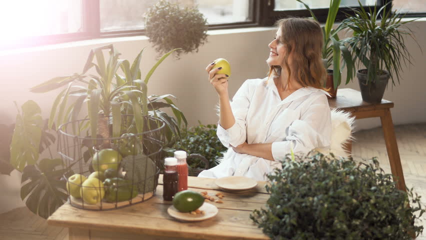 Fair-haired girl smelling a green apple with pleasure at the table full of healthy food, indoor shot among green plants