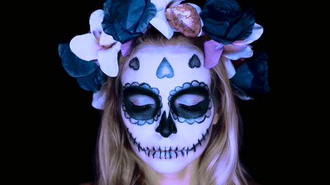 Closeup face of woman with Mexican sugar skull makeup and flowery wreath opening eyes and looking into the camera. Creative, artistic, Halloween concept - slow motion video