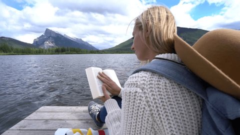 Young woman sitting on wooden pier reads book.
Female relaxing on wooden jetty by the lake, reads a book surrounded by nature 