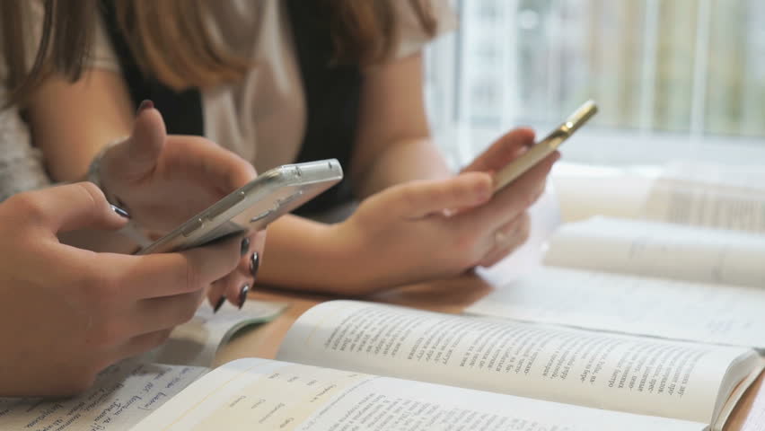 Two unidentified female students sit at wooden desk at lesson at the university and thumb their fingers on the smartphone