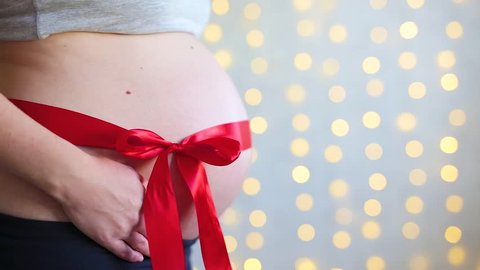 pregnancy and christmas concept - expectant mother with red ribbon on her belly and male hands unleashing a bow