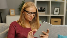 Beautiful young blonde woman in glasses using the tablet on the sofa in the cosy living room. Portrait. Indoor