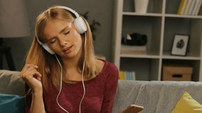 Charming blonde young woman enjoying the music through the white headphones on the smart phone, moving to the rhythm in the comfortable living room. Indoor