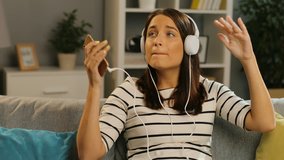 Attractive young woman in the white headphones listening to the music on the smart phone, singing and dancing in the nice living room. Indoors