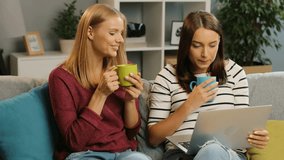 Two beautiful young women talking, drinking tea and looking at the laptop screen on the sofa in the cosy living room atmosphere. Home